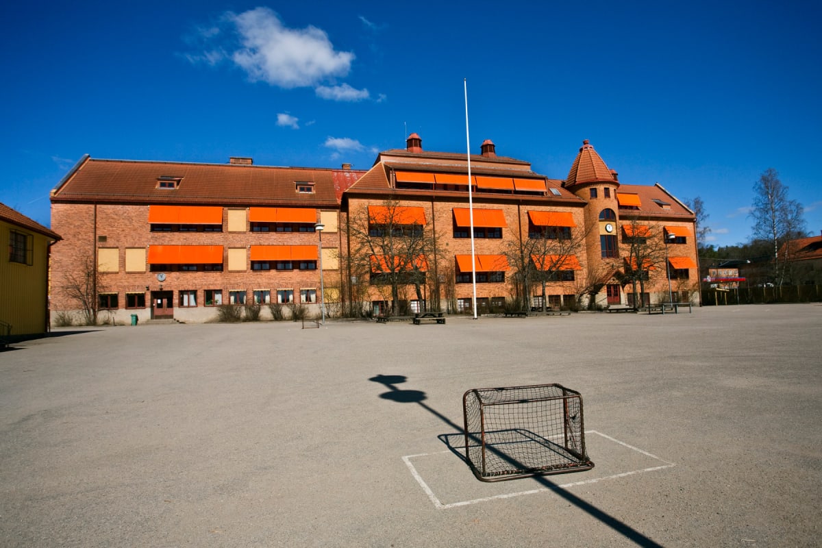 En stor röd tegelbyggnad med orangefärgade fönstermarkiser under en klarblå himmel med ett vitt moln. Framför byggnaden ligger en asfalterad skolgård med en liten fotbollsmålbur och en flaggstång skugga på marken.