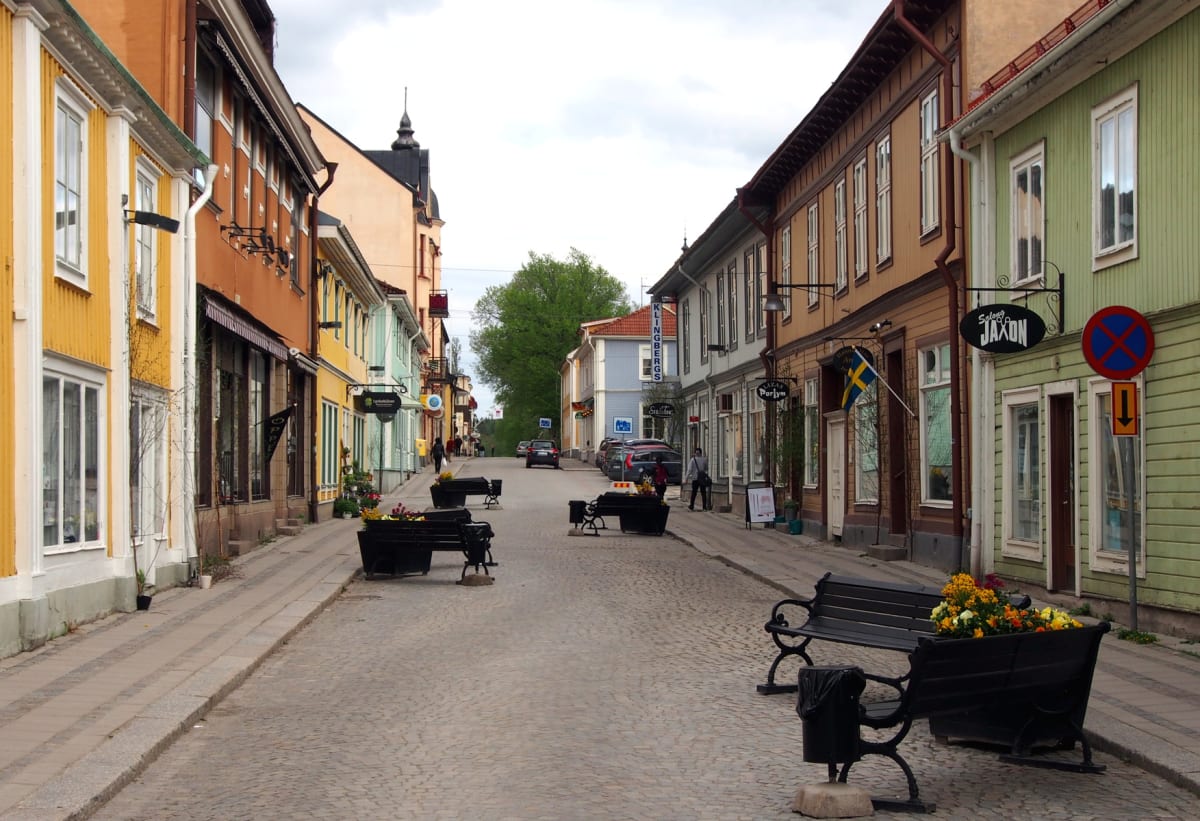 En gata med gatsten belagd och kantad av flervåningshus i olika färger, inklusive gula, orangea och mintgröna fasader. Längs gatan står bänkar med blomlådor och det syns människor, parkerade bilar och en svensk flagga. Ett "parkering förbjuden"-skylt