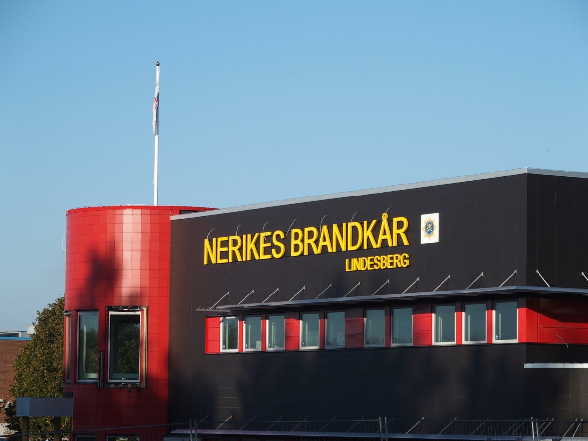 En modern byggnad med röd och svart fasad. På den svarta delen syns en skylt med Gul text ”NERIKES BRANDKÅR” och undertill ”LINDESBERG” samt en logotyp. Ett flaggstång med en flagga syns på taket. Fönster syns längs fasaden. Ett lövträd står intill b