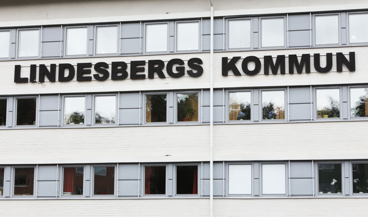 En fasad på en byggnad i ljus tegel syns i närbild. Texten på fasaden identifierar byggnaden som "LINDESBERGS KOMMUN". Fönster med gråa ramar är synliga, och i några av dem syns reflektioner av grönska samt inomhusväxter.