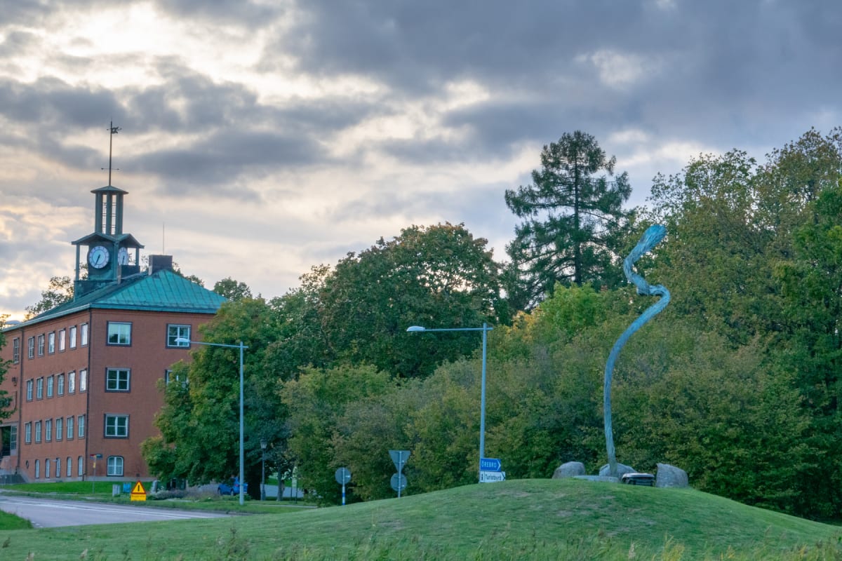 En bild visar en tegelbyggnad med ett klocktorn och en grön koppartak under en mulen himmel. Framför byggnaden finns ett grönområde med träd, en vägbana, och en blå skylt som pekar mot 'ÖREBRO' och 'Turistbyrå'. Till höger i bilden står en blå-grön s