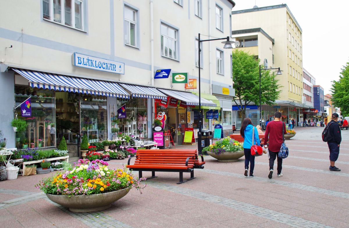 En gata med butiker, människor och blomsterarrangemang. På vänster sida av bilden syns en butiksskylt med texten "BLÅKLOCKAN" och en markis med blåvita ränder. Fasaderna på byggnaderna är ljusa, och gatan är asfalterad med inslag av grå marksten. Fle
