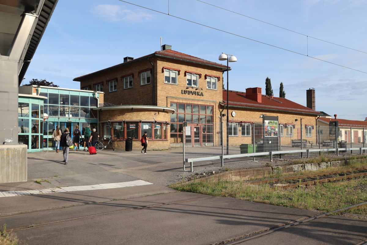 En bild som visar Ludvika station med byggnad i tegel och ett rött tak. Framför stationen finns en asfalterad yta med människor och en cykel. Tågspår löper i förgrunden och strömkablar syns i den blå himlen.