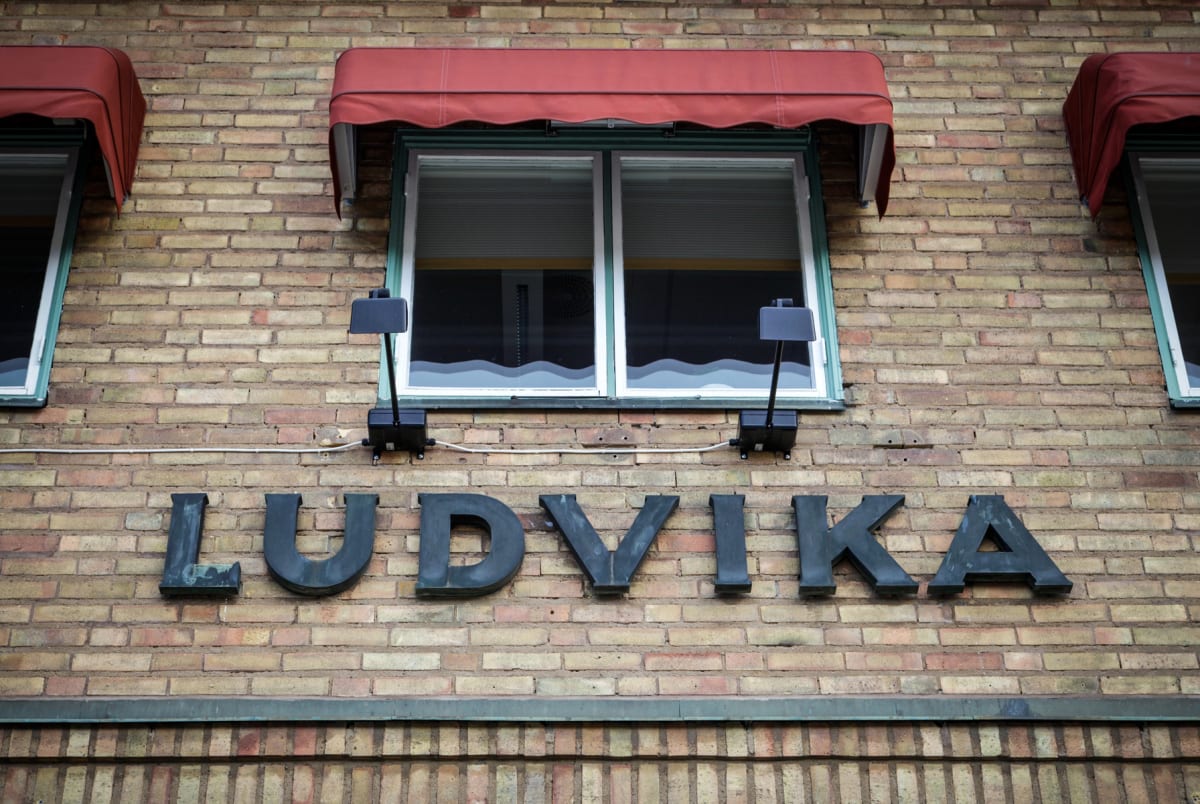 En fasad av tegel med texten "LUDVIKA" monterad undertill. Ovanför texten finns ett stort fönster med vita karmar och två mindre fönster med röda markiser. Två svarta justerbara lampor är fästade på fasaden under det stora fönstret.