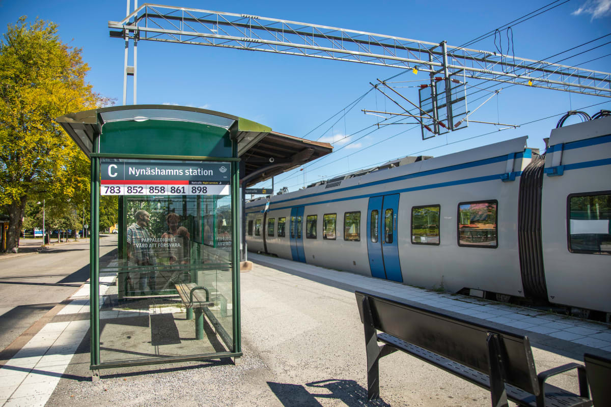 En bild som visar en tågstation med ett tåg på spåret, en entré till en busskur eller vänthall med skylten "Nynäshamns station", samt gult lövverk på träd i bakgrunden. En solig himmel syns ovanför.