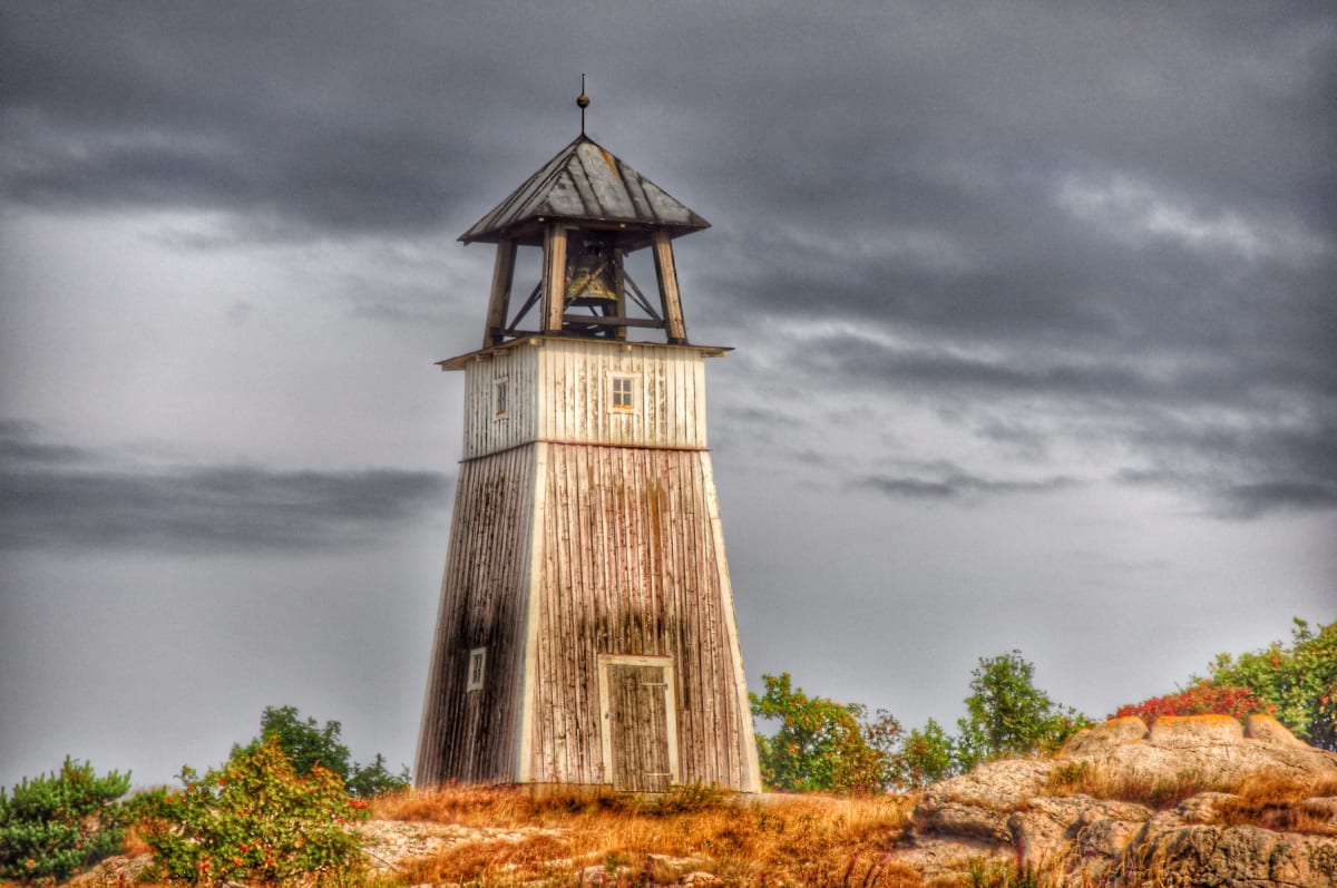 En stor, vit träbyggnad med en klocka i toppen under en mörk och mulen himmel. Byggnaden står på en klippig höjd med torrt gräs och buskar med både gröna och röda löv. Klockan syns tydligt i öppningen av byggnadens övre del.