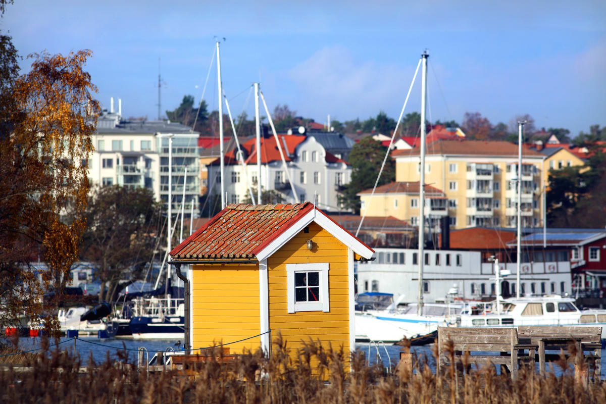 En gul byggnad med rött tak och ett vitt fönster står i förgrunden, omgiven av vass. Bortom byggnaden syns ett vattendrag med flera båtar och segelbåtar. I bakgrunden syns bostadshus med fasader i olika färger samt träd med höstlövsfärg. En bänk med