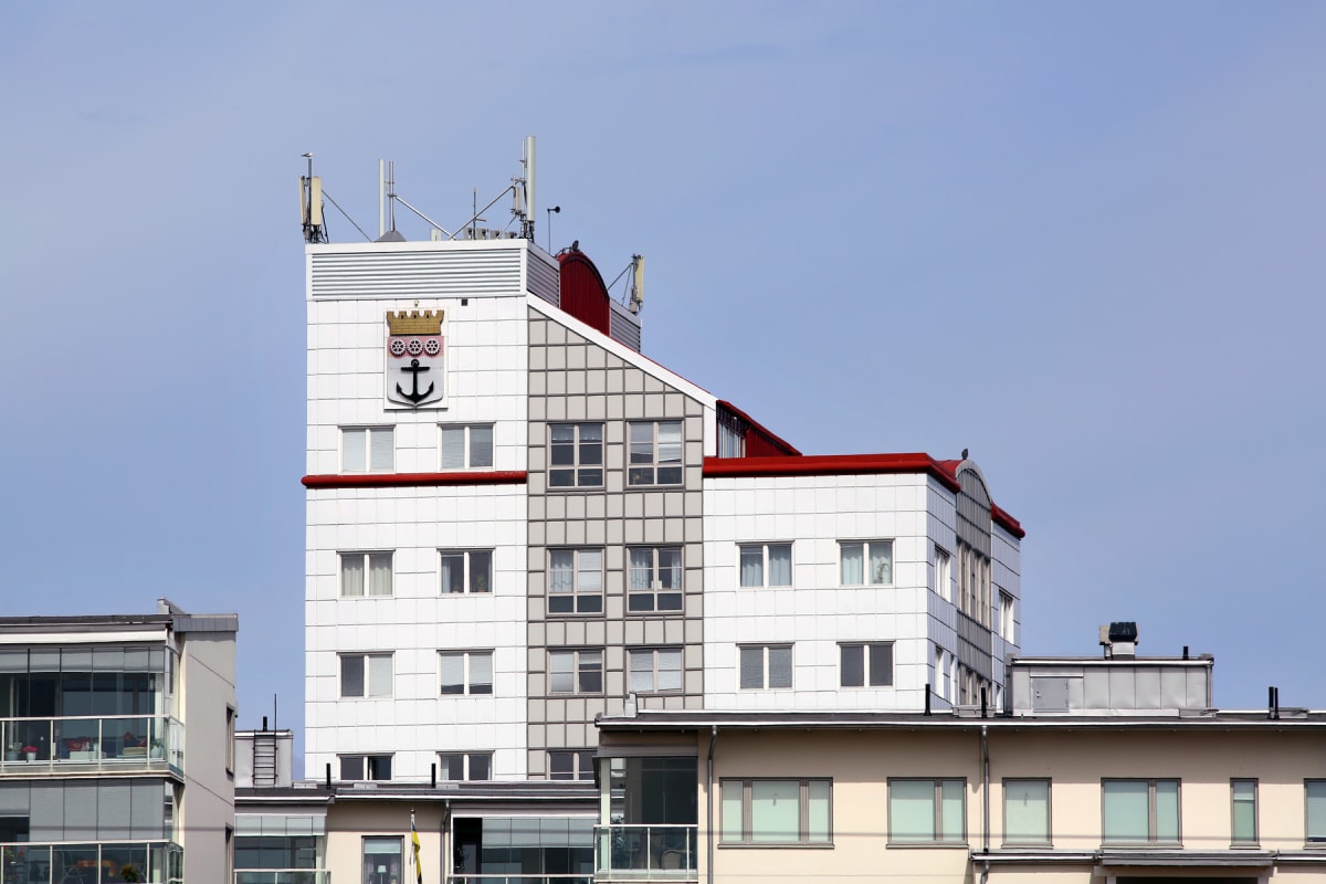 En modern byggnad med vita och grå fasader samt röda takdetaljer. På byggnadens front finns en vapensköld med en ankare omgiven av kugghjul och en krona. Flera fönster syns, samt antenner på taket.