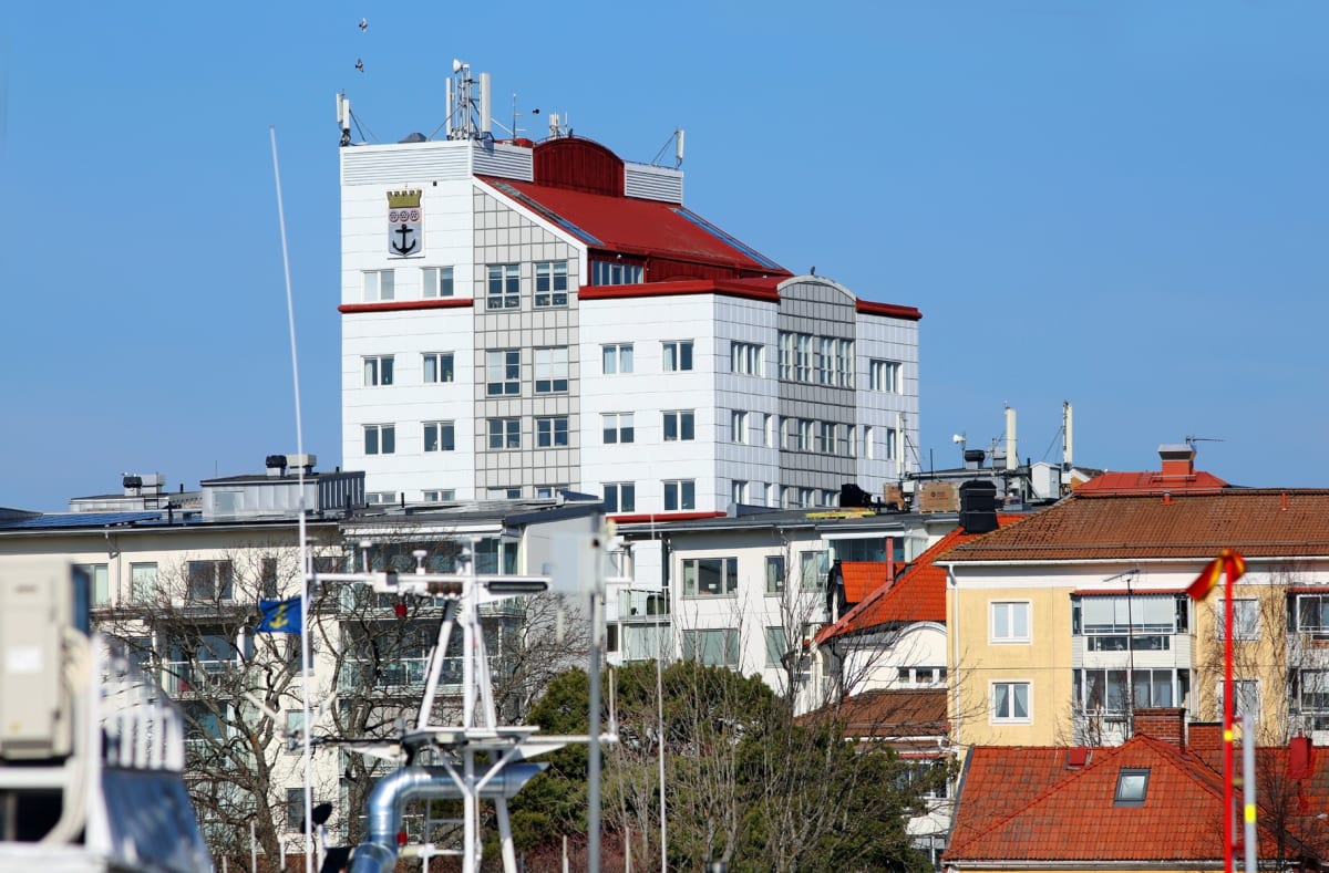 En blandning av byggnader i ett stadslandskap med en stor vit byggnad i centrum med ett rött tak och en vapensköld. Det är en klarblå himmel med några fåglar synliga. På flera tak syns olika installationer.