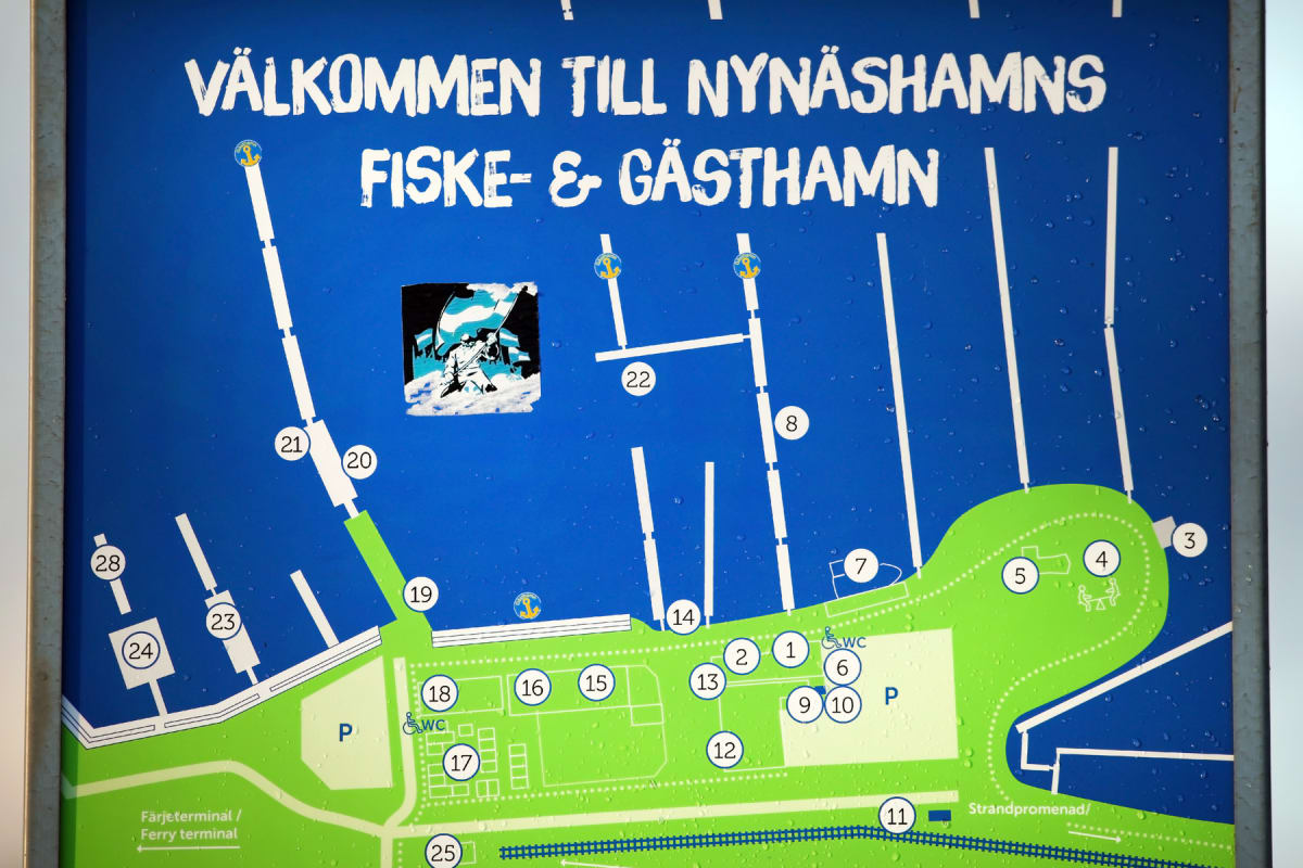 En väggmonterad blå och grön karta över Nynäshamns fiske- och gästhamn, med texten "VÄLKOMMEN TILL NYNÄSHAMNS FISKE- & GÄSTHAMN" överst. Kartan visar en hamn med båtplatser, byggnader, parkeringsplatser och en strandpromenad. Vattendroppar täcker kar