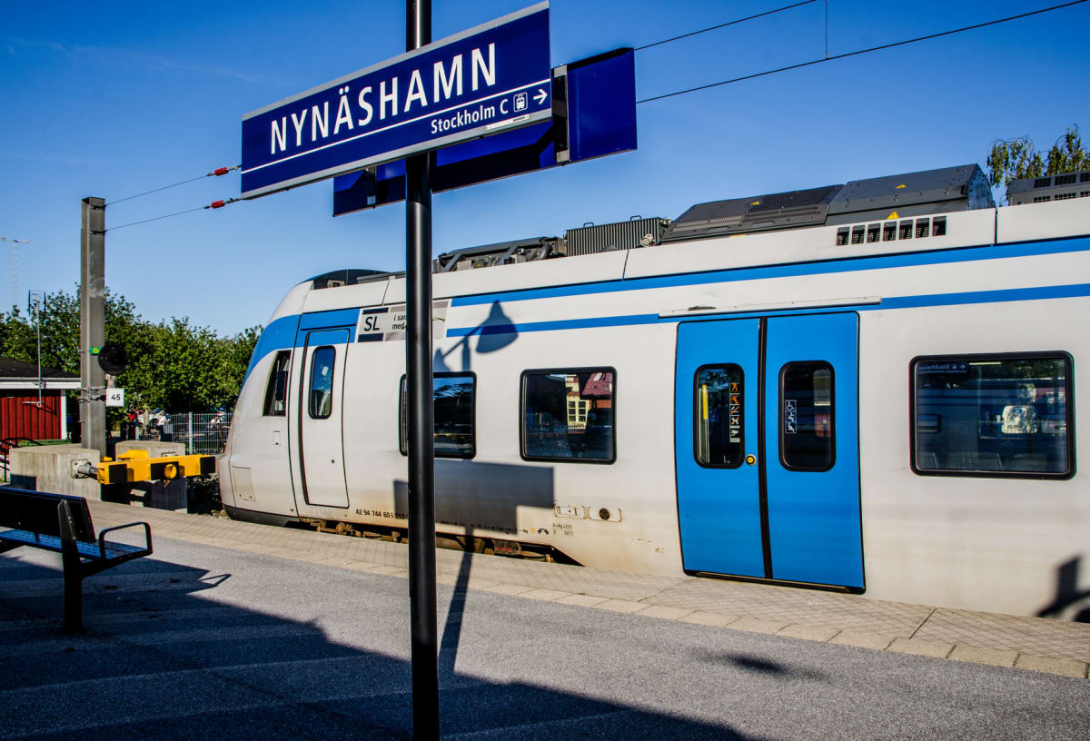 En bild tagen utomhus i dagsljus som visar den mittersta delen av ett vitt och blått tåg som står på en perrong. En blå skylt med texten ”NYNÄSHAMN” och en pil mot ”Stockholm C” syns tydligt i förgrunden. En svart bänk syns i nedre vänstra hörnet, oc