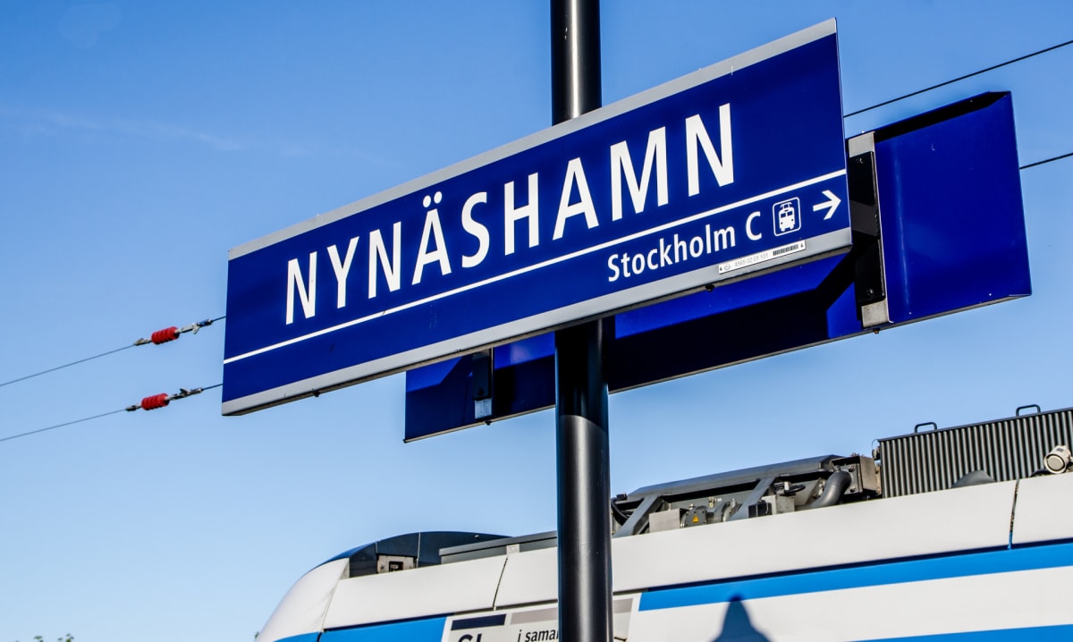En blå skylt med texten "NYNÄSHAMN" och "Stockholm C" samt en riktningspil och tågsymbol, monterad på en svart stolpe. En del av ett vitt och blått tåg är synligt i bildens nedre del. Himlen är klarblå och ljus.