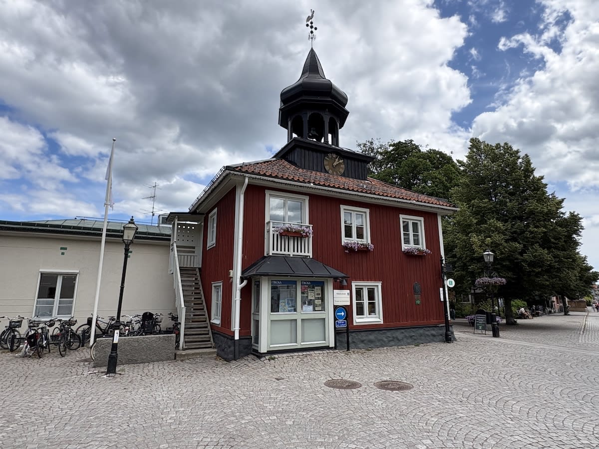 En röd träbyggnad med vit spröjsade fönster och en svart takspira med en vindflöjel. Till vänster om byggnaden syns en trappa som leder upp till en dörr och längre bort en ljusare byggnad. Ett antal cyklar är parkerade vid den ljusare byggnaden. Fram