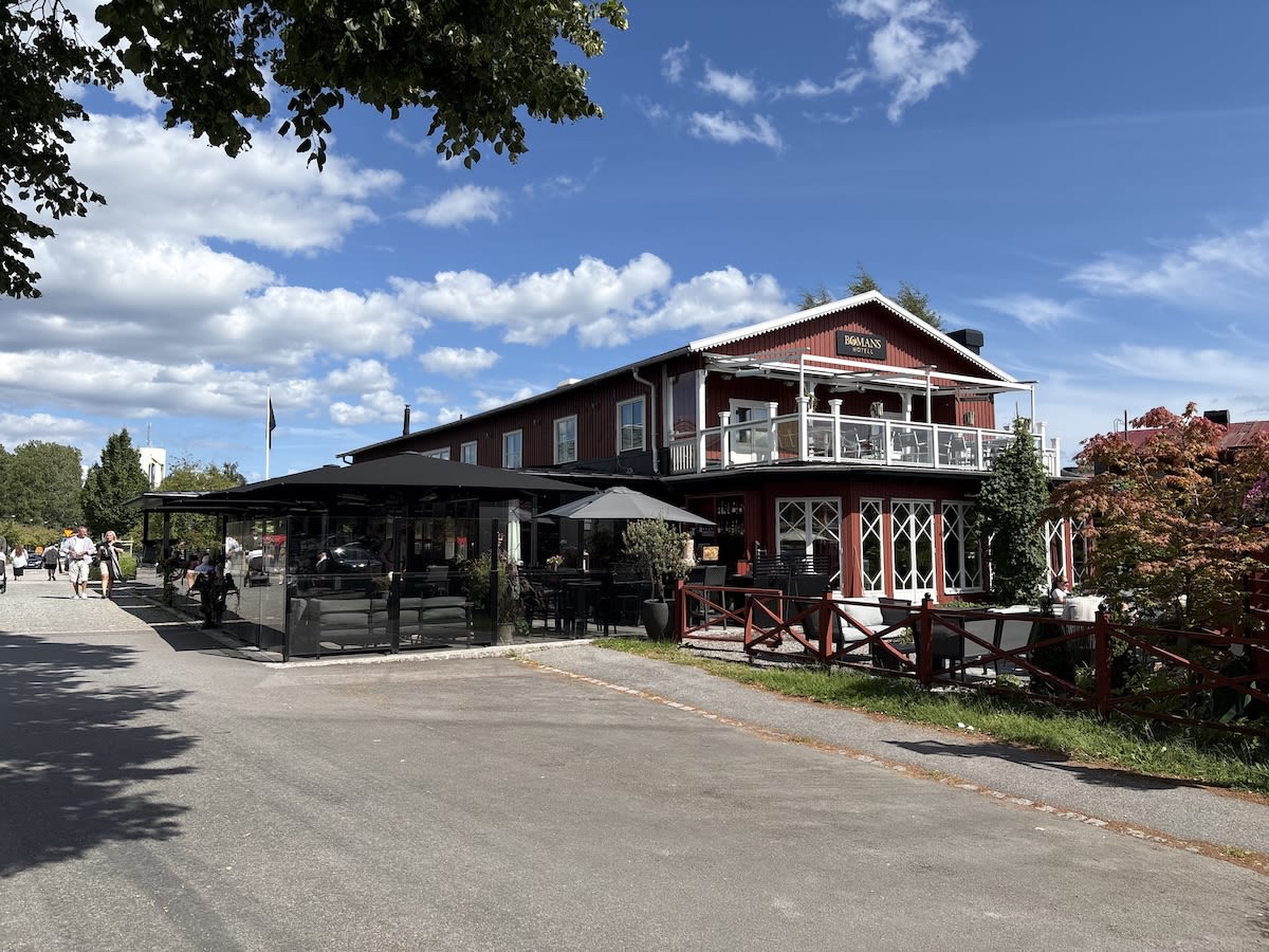 En röd byggnad med vita knutar och en balkong syns under en blå himmel med moln. Framför byggnaden finns en uteservering med svarta tak och glasväggar, samt en grusgång där människor promenerar. Gröna träd och buskar ramar in bilden.