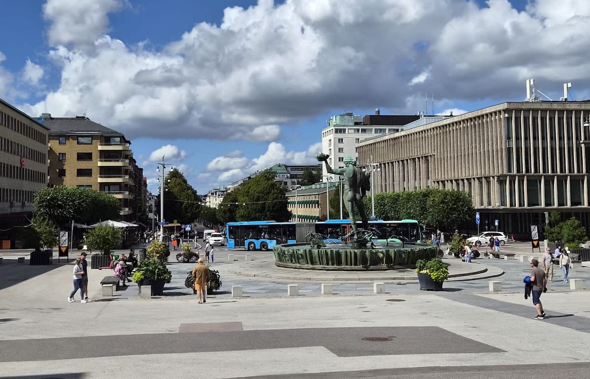 En vidvinkelbild tagen utomhus under dagtid, som visar en staty föreställande Poseidon vid en fontän, omgiven av människor, bussar och byggnader under en delvis molnig himmel. Gatan är trafikerad och det finns grönska i form av träd och buskar.