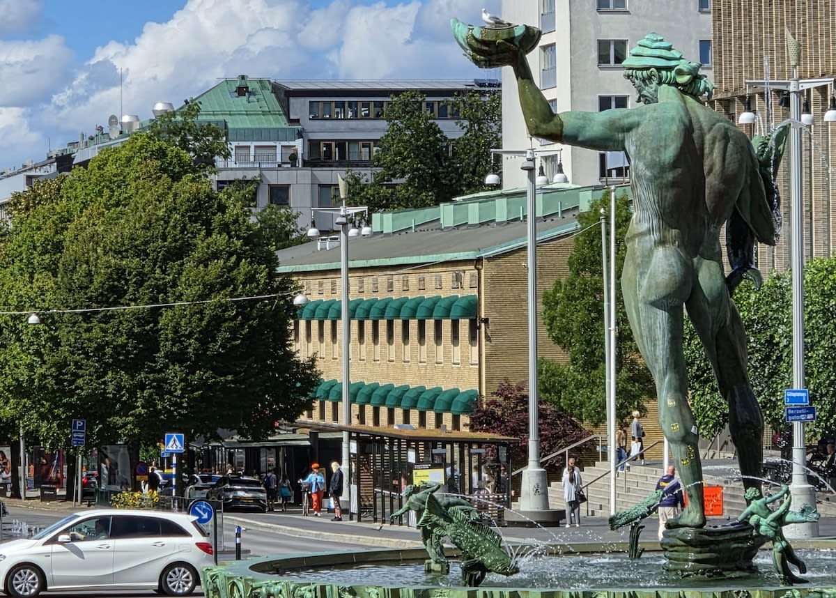 En stor bronsstaty föreställande en man som håller en stor skål i sin uppsträckta hand, där en mås sitter. Statyn är en del av en fontän med vatten som sprutar ut. I bakgrunden syns byggnader, träd, en väg med fordon och människor.
