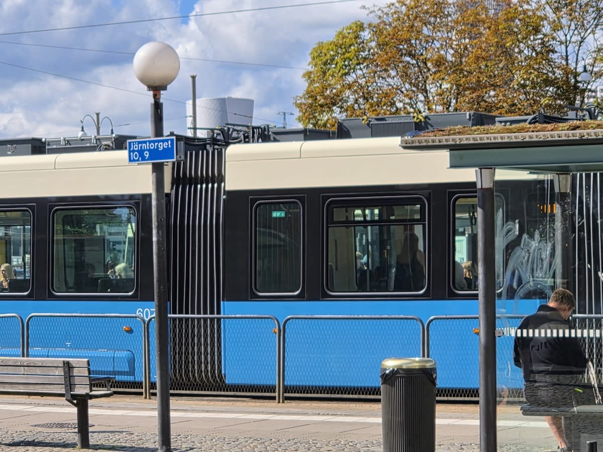 En spårvagn i blått och vitt står vid en hållplats med en bänk och en papperskorg. En skylt med texten "Järntorget 10, 9" syns på en stolpe. Träd med vissna löv syns i bakgrunden, och en person sitter på en bänk bakom en glasvägg.