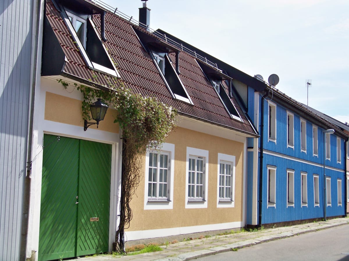 En bild som visar en rad hus utomhus en solig dag. Det första huset är ljust gult med grön dörr, vita fönsterkarmar och brunt tak. Ett blått hus syns bredvid, också med vita fönsterkarmar. Klätterväxter växer på det gula husets fasad.