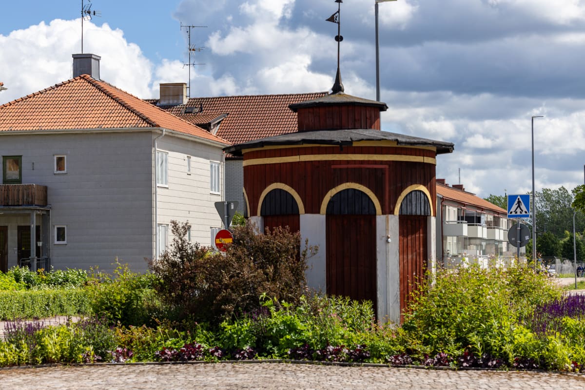 En bild som visar en rödbrun, åttakantig byggnad med svarta bågformade fönster och en svart takkupol med en flagga på toppen. Bakom syns bostadshus med tegeltak och antenner. Grönskande buskar och blommor finns i förgrunden.