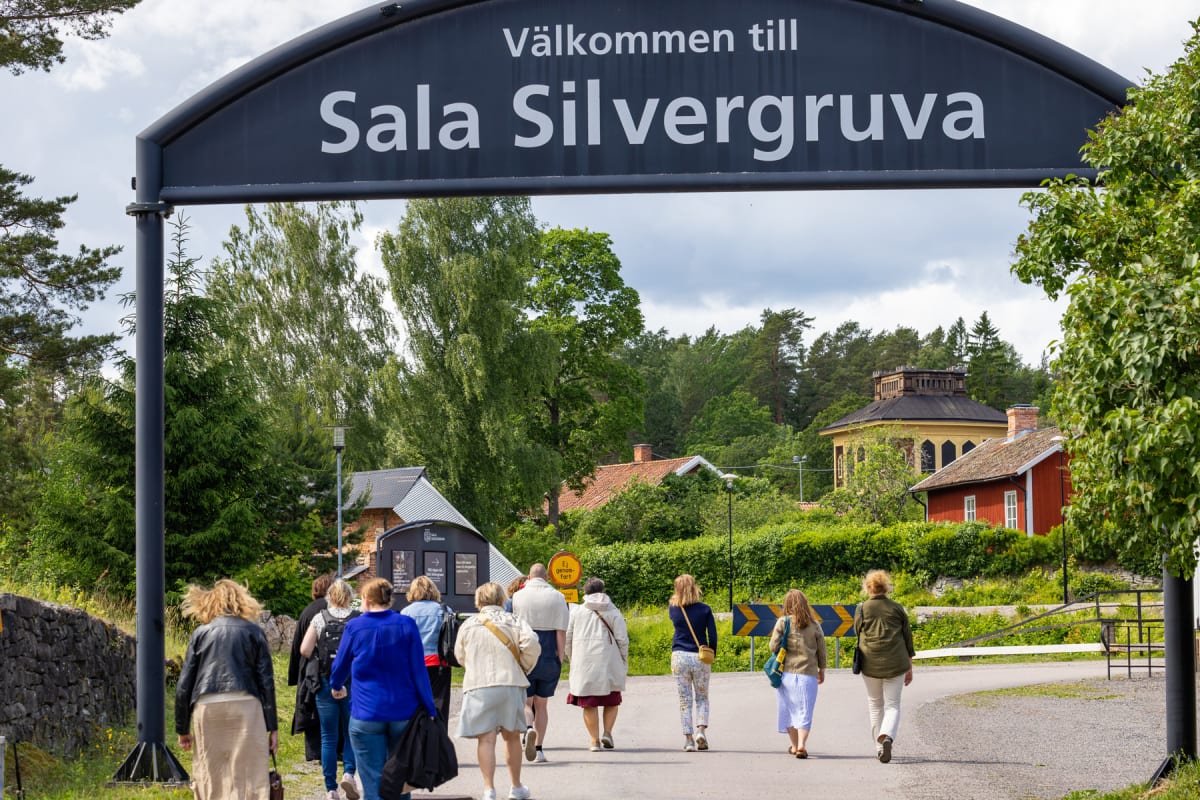 En grupp människor går på en väg under en stor skylt med texten "Välkommen till Sala Silvergruva". I bakgrunden syns grönskande träd och byggnader, inklusive ett gult hus med torn och ett rött hus med vita knutar.