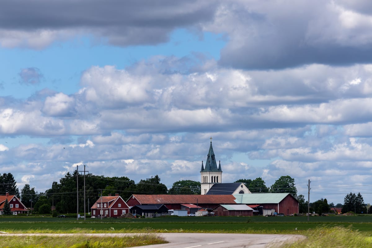 En bild som visar en kyrka med spira i bakgrunden, omgiven av röda byggnader med lador. I förgrunden syns en bit av en väg och ett grönt fält. Himlen är täckt av moln, med en del blå himmel synlig.