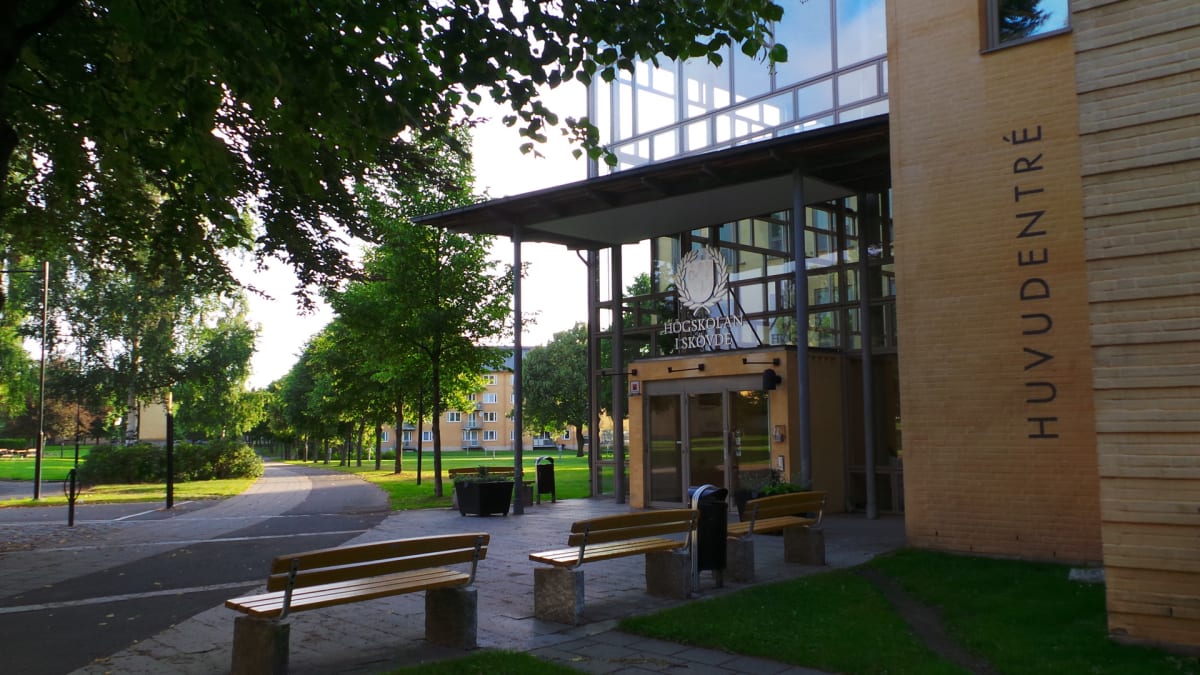 En bild av huvudentrén till en byggnad märkt "HÖGSKOLAN I SKÖVDE" och "HUVUDENTRÉ" på en solig dag. Flera gröna träd, bänkar och en gångväg syns framför byggnaden. Ett bostadshus med gula fasader skymtar i bakgrunden till vänster.