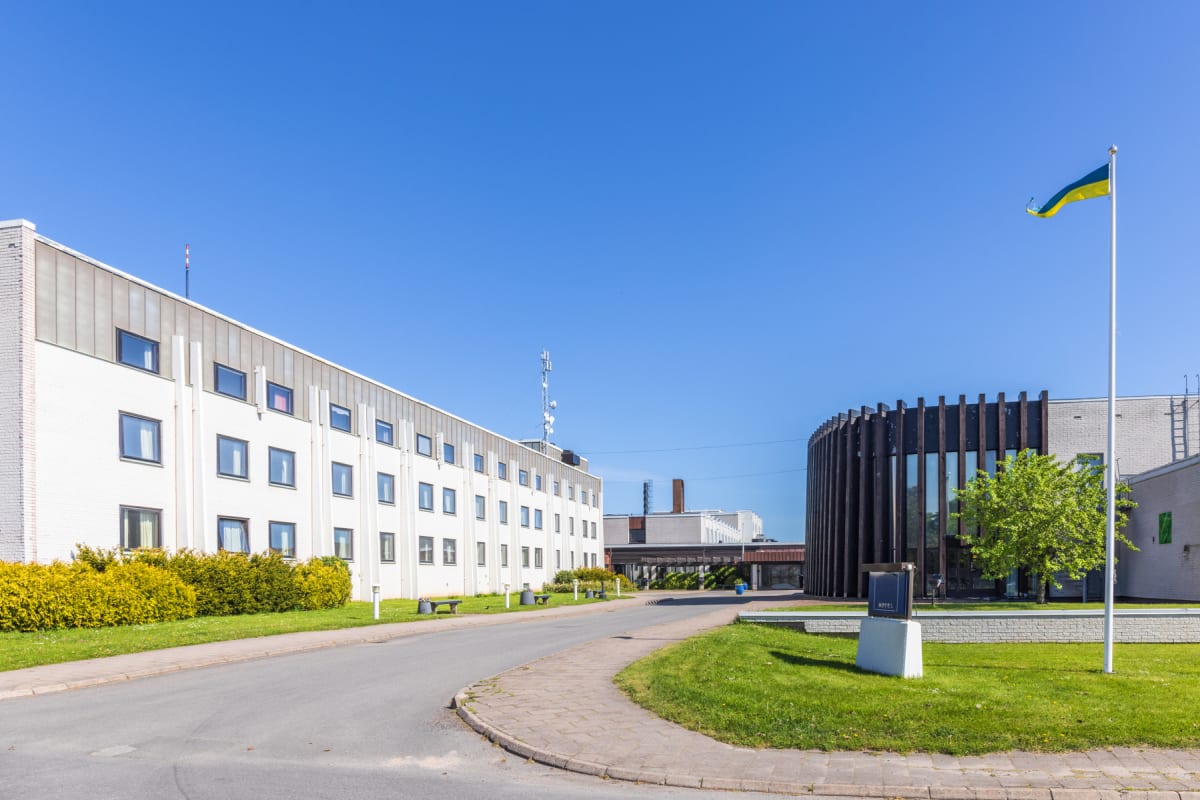 En utomhusbild som visar byggnader med en flaggstång och en vajande flagga i färgerna blått och gult. Mellan byggnaderna finns gräsmattor, buskar och en asfalterad väg. En av byggnaderna är vit med många fönster, och den andra har en rundad fasad med