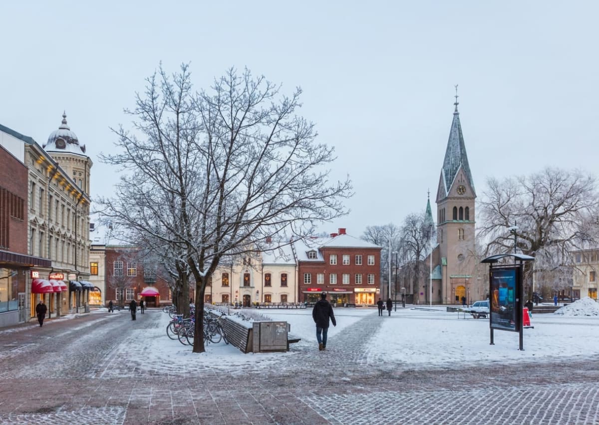 Bilden visar ett snötäckt torg med byggnader runt omkring under dagtid. Flera personer promenerar på gångbanorna och det syns träd, varav ett stort i förgrunden är täckt av snö. Till höger reser sig en kyrka med ett högt torn, och flera butiksfönster