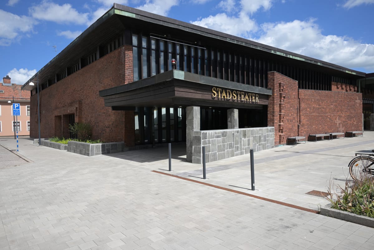 En bild av en byggnad med texten "STADSTEATER" på en träfasad. Byggnaden har tegelväggar och stora fönsterpartier. Framför byggnaden finns ett stenbelagt torg med planteringar, sittbänkar och pollare. En parkeringsskylt och en cykel syns i bildens ka