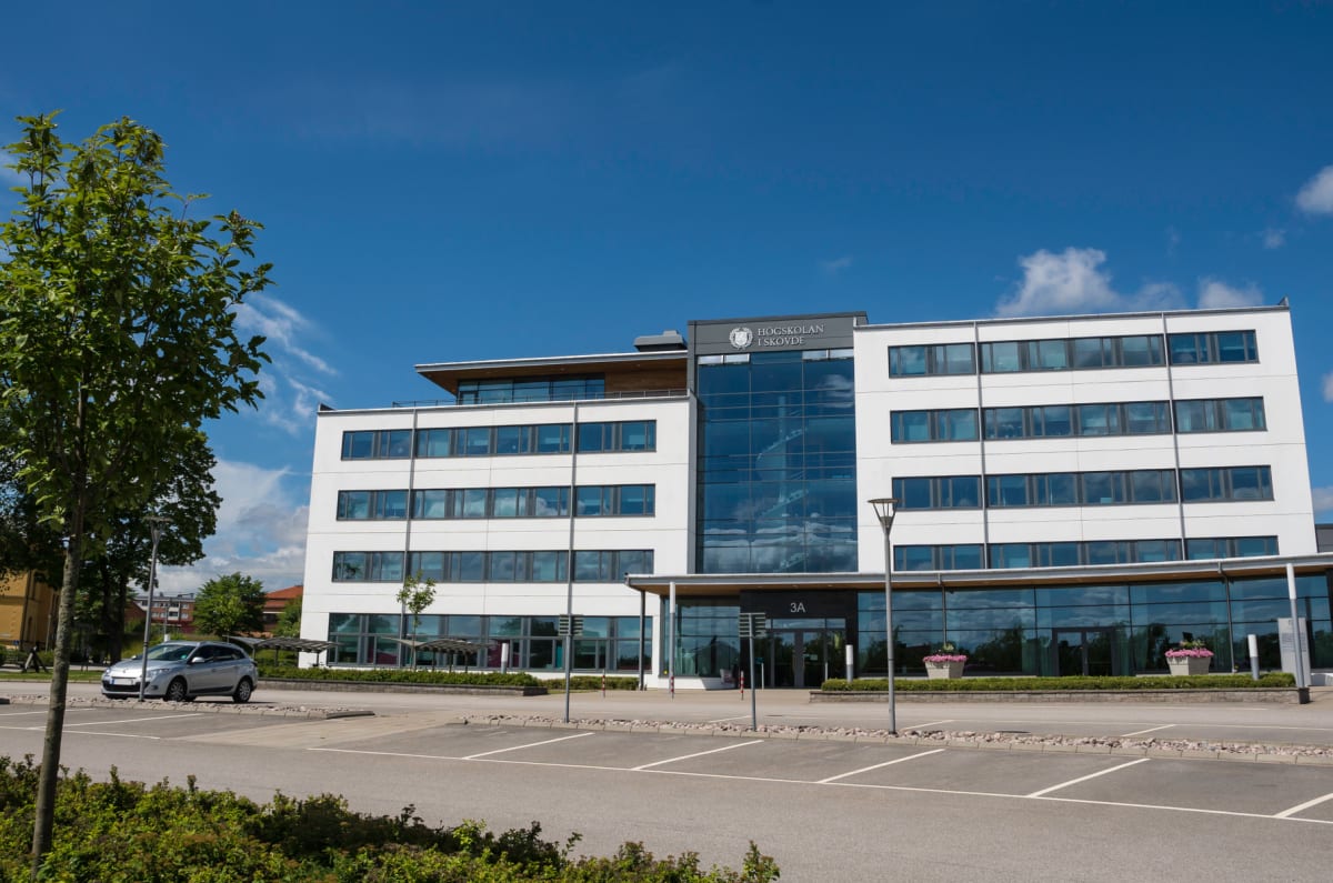 En modern byggnad med stora fönster och en logotyp som lyder "HÖGSKOLAN I SKÖVDE" under en blå himmel. Framför byggnaden finns en parkeringsplats med en parkerad silverfärgad bil, samt grönskande buskar och ett träd till vänster.