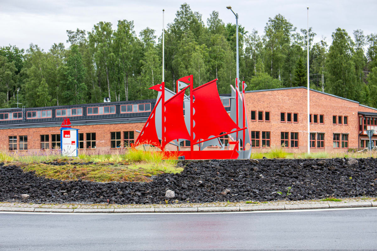 En stor, röd och grå skulptur föreställande ett fartyg står på en gräsbevuxen kulle omgiven av mörkgrå sten. Bakom skulpturen syns en tegelfärgad byggnad med många fönster och en skylt. I bakgrunden finns en grön skog och en grå himmel. Asfaltväg och
