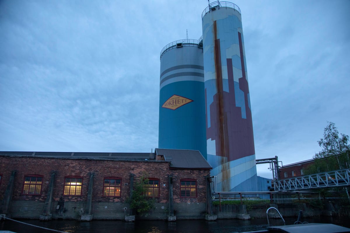 En bild som visar en tegelbyggnad vid vatten, med upplysta fönster, och bakom den reser sig två stora silos mot en molnig himmel. En av silon har texten "KOKHETT" och "FJÄRRVÄRME" på en guldfärgad rombformad skylt.