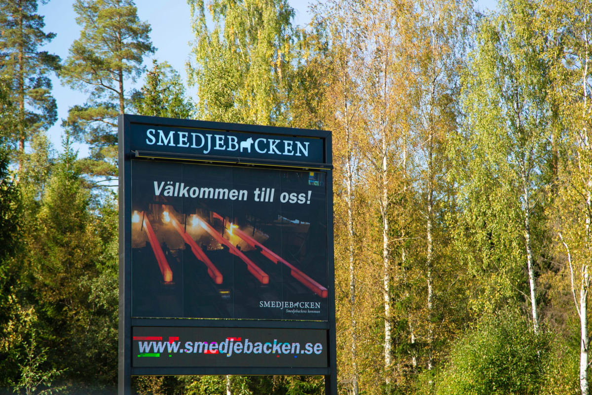 En vertikal bild av en digital informationsskylt med texten "SMEDJEBACKEN", "Välkommen till oss!" och "www.smedjebacken.se". Bakgrunden är skog med träd som visar höstfärger, där björkar med gula och gröna löv dominerar.