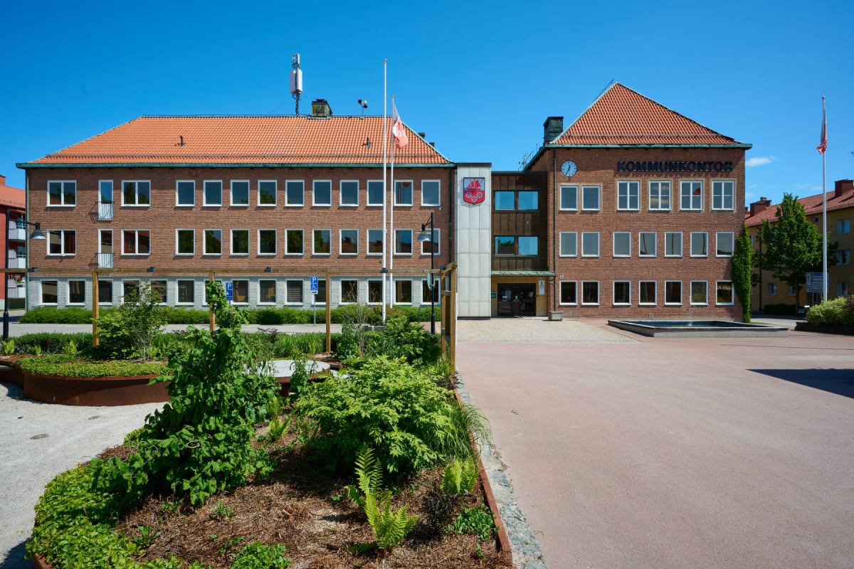 En stor, röd tegelbyggnad med tegeltak och många fönster dominerar bilden under en klarblå himmel. Framför byggnaden finns ett öppet torg med en fontän, en flaggstång med en flagga, samt planteringar med gröna buskar och växter. På byggnadens fasad s