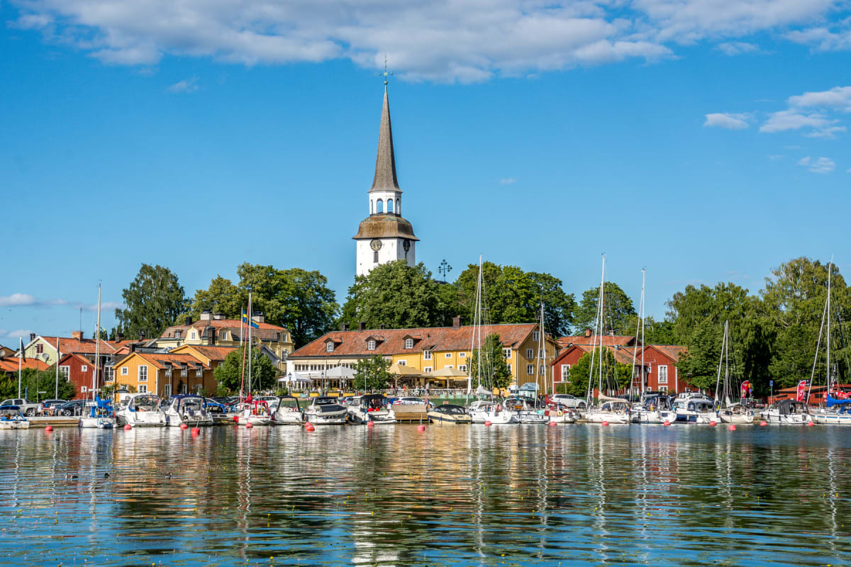 En bild tagen över vatten som visar en hamn med många båtar förtöjda längs kajen. Bakom båtarna syns en rad med byggnader i olika färger, samt en vit kyrka med en hög spira. Himlen är blå med några vita moln och träden är gröna.