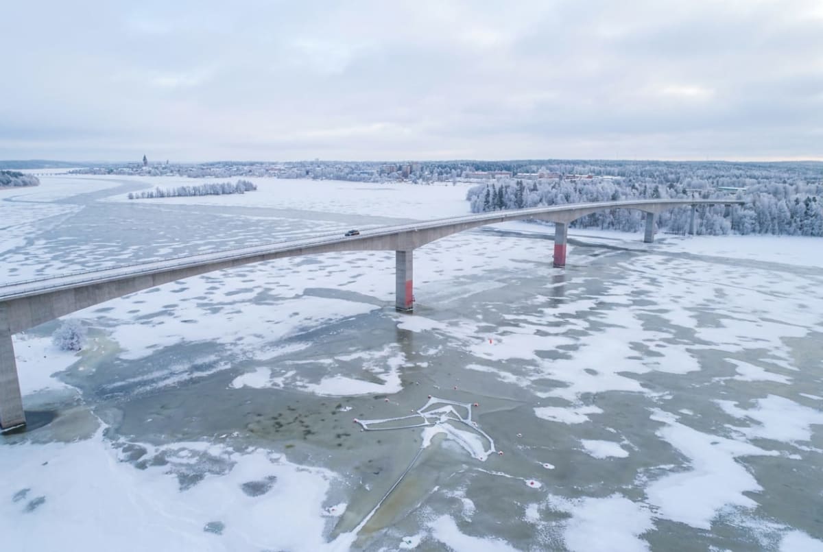 En vidvinkelbild från ovan visar en bro som sträcker sig över ett is- och snötäckt vatten. På bron syns ett fordon, och i bakgrunden finns en stad med byggnader och träd. Landskapet är täckt av snö och is, vilket indikerar vinter.