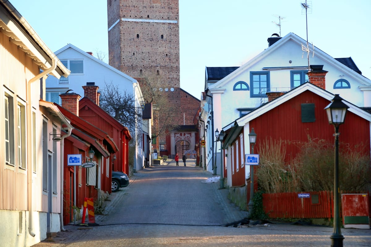 En gata kantad av hus med röda och vita fasader, där en stor tegeltornbyggnad syns i bakgrunden. Två personer står längst ner på gatan och en bil är delvis synlig parkerad på vänster sida. Gatlyktor och skyltar syns längs gatan.