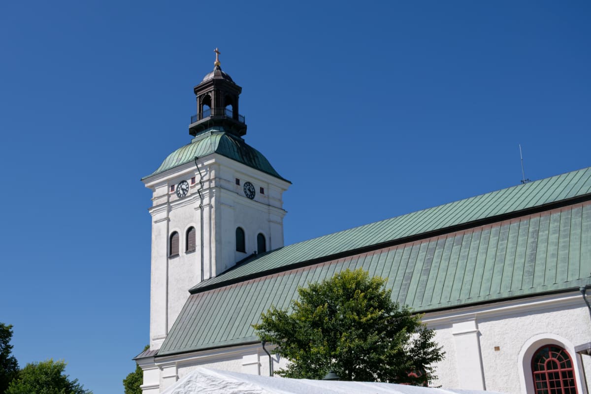 En vit kyrka med grönt tak och ett klocktorn syns mot en klarblå himmel. Delar av gröna träd och en vit tältstruktur syns i förgrunden.