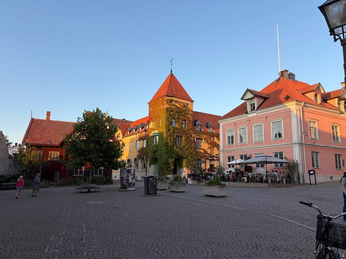 En bild av ett torg eller en öppen plats med kullersten framför flera byggnader i olika färger, inklusive en rosa byggnad och en gul byggnad med klätterväxter. Det finns en uteservering med parasoller och folk som sitter vid bord. Två personer står p