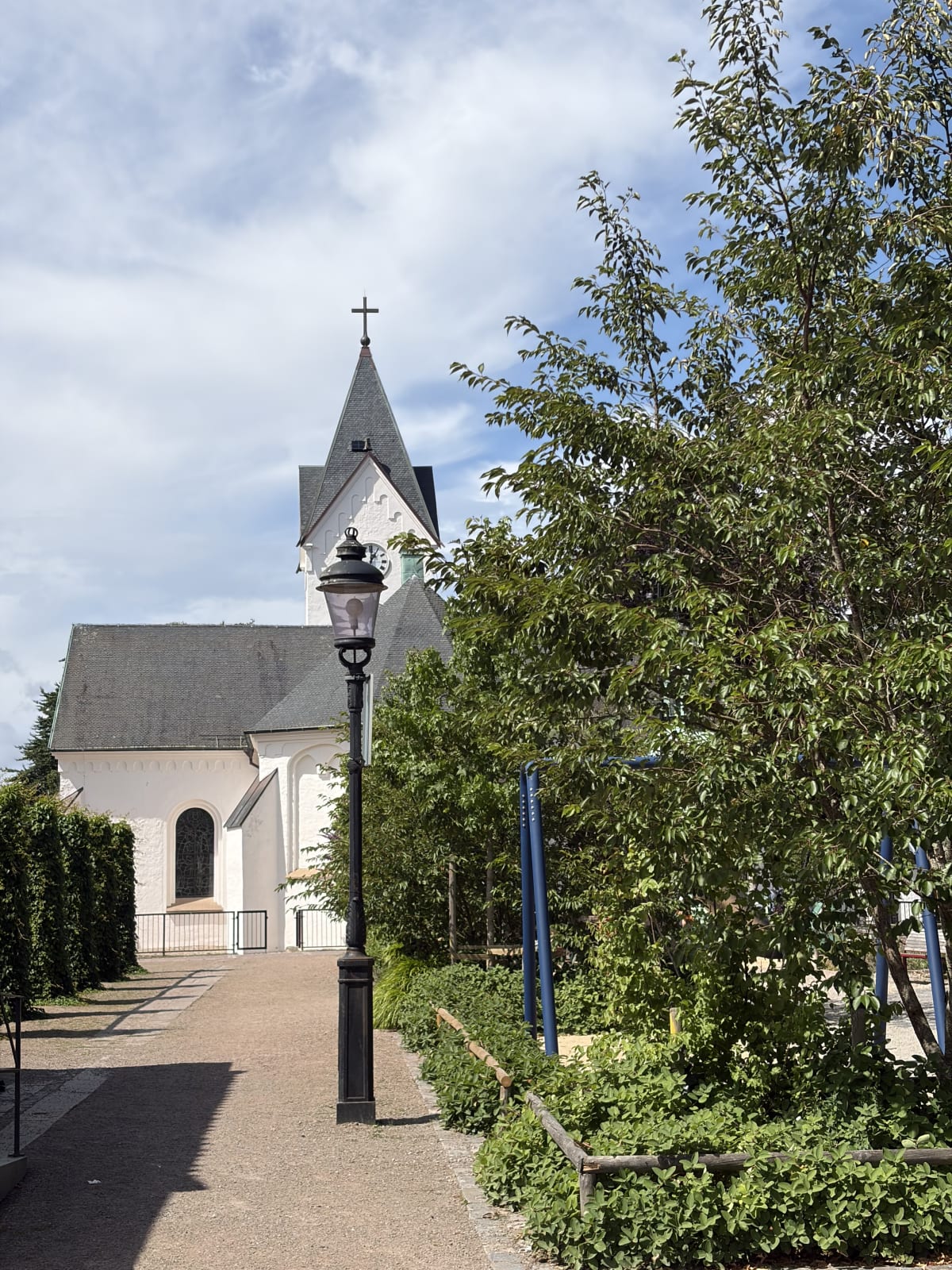En bild som visar en vit kyrka med ett grått tak och ett kors på toppen av spiran, omgiven av grönska och träd. I förgrunden syns en lyktstolpe och en grusgång. En blå stång är också synlig i bilden. Ängelholms kyrka.