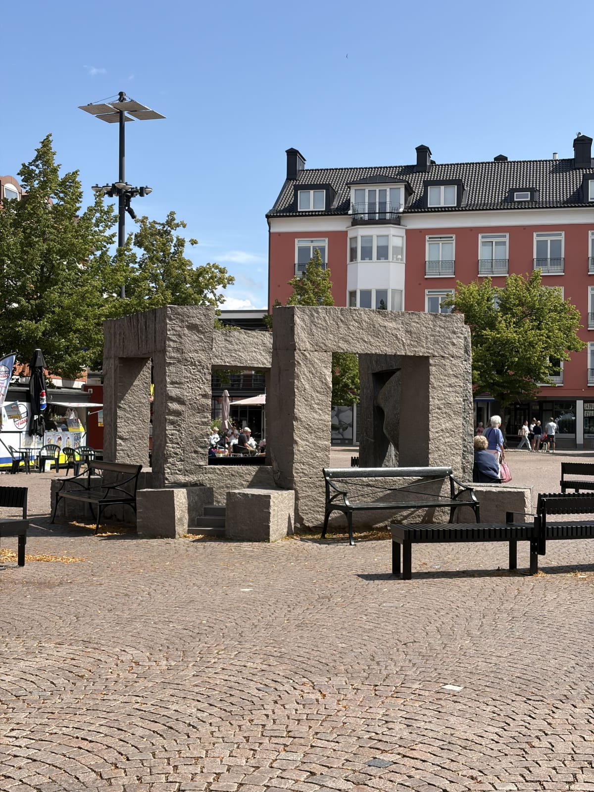 En bild av ett torg eller en stadsmiljö med en stor stenkonstruktion i förgrunden, omgiven av gatsten. I bakgrunden syns ett rött flerfamiljshus och ett grönt träd. Flera människor syns sittandes på bänkar och vid ett utecafé.