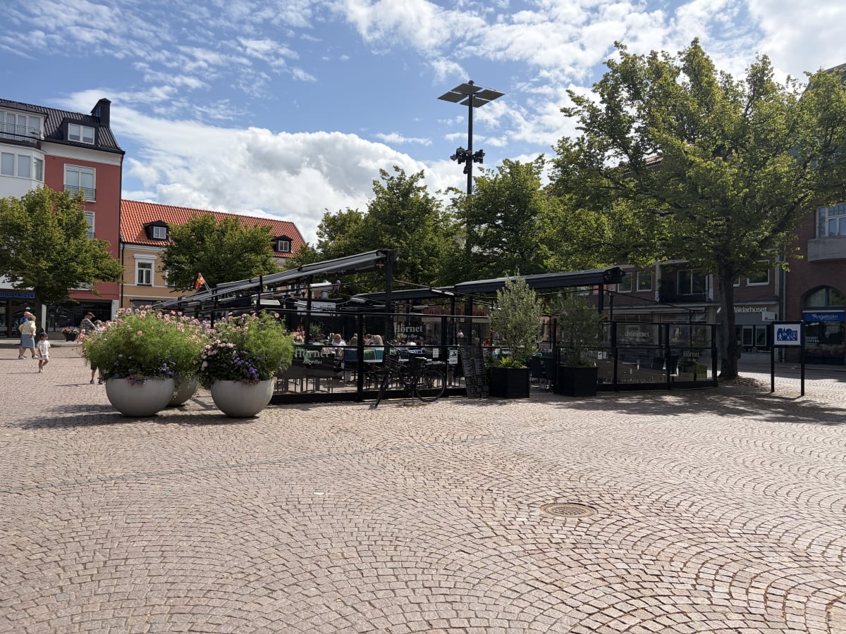 En utomhusbild som visar en kullerstensbelagd plats med ett uteserveringsområde omgivet av glasväggar. Flera träd med grönt lövverk och byggnader med röda tak syns i bakgrunden. Tre stora krukor med blommor står i förgrunden.