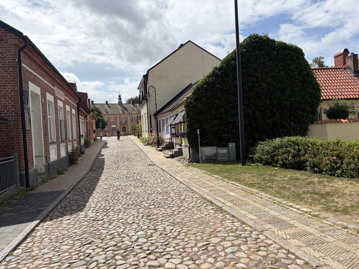 En kullerstensgata med byggnader på båda sidor under en molnig himmel. På vänster sida syns röda tegelhus och på höger sida ett ljust hus delvis täckt av murgröna. Två personer är synliga längre bort på gatan, varav en cyklar.