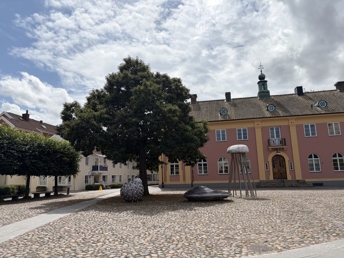 En bild av ett torg med kullersten framför en rosa byggnad med gula detaljer och ett grått tak. På torget står en stor grönlummig träd, samt flera skulpturer, varav en metallisk klotformad och en metallisk med tentakelliknande utskott. En liten del a