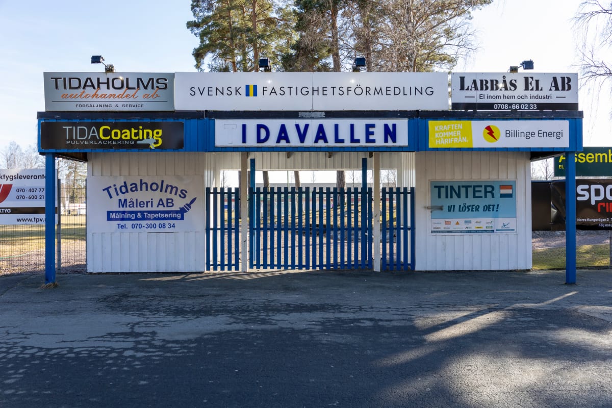 En entrébyggnad till 'TIDAVALLEN' med blåa grindar och flera reklamskyltar för lokala företag. Solen skiner och träden i bakgrunden har inga löv, vilket indikerar tidig vår eller sen höst.
