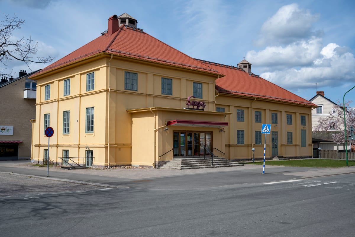 En gul byggnad med rött tak och texten "Saga" ovanför huvudentrén. Framför byggnaden finns en asfalterad väg, en gångbana, samt ett övergångsställtecken och ett stoppskylt. Molnig himmel och grönska syns i bakgrunden.