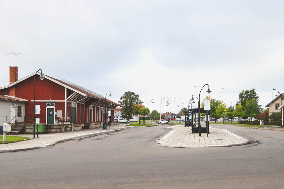 En gata med en röd träbyggnad till vänster och en busshållplats till höger. Träd med grönskande löv syns i bakgrunden. Ett flertal flaggstänger med vita flaggor står uppställda. På gatan finns även en grön soptunna och ett par bänkar utanför byggnade