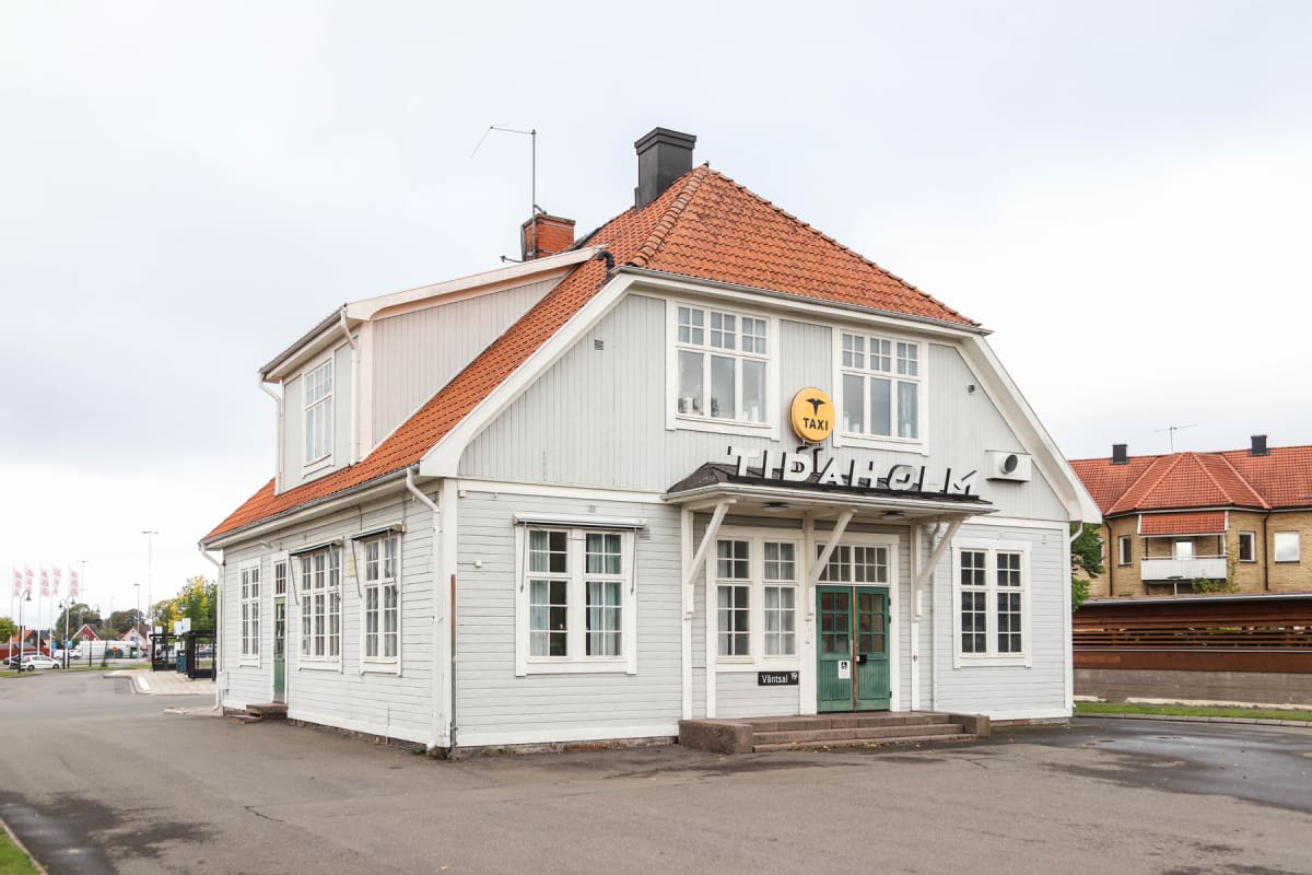 En ljusgrå byggnad med orange takpannor står centralt i bilden. På byggnadens framsida sitter en gul skylt med texten "TAXI" och en kråka, samt bokstäverna "TIDAHOLM" ovanför entrén. En grön dörr och flera fönster är synliga. Asfalt och gräs syns i f