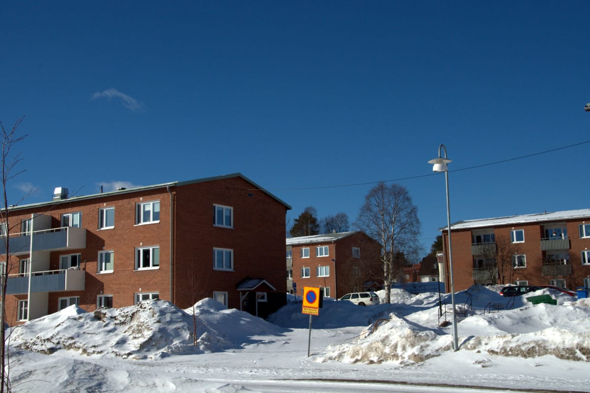 En gata täckt av snö framför flera röda tegelhus under en klarblå himmel. Stora snöhögar syns längs sidorna av vägen, och en gatlykta står i förgrunden. En parkeringsskylt med gul botten och blå text är synlig. Träd utan löv står mellan byggnaderna.