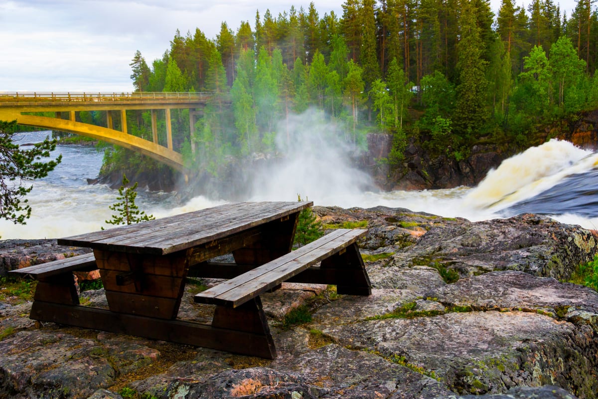 En bild som visar en fors med en bro i bakgrunden, omgiven av tallskog. I förgrunden syns ett träbord med bänkar på en stenig mark. Vattenånga stiger från forsen. Himmeln är molnig och grå.
